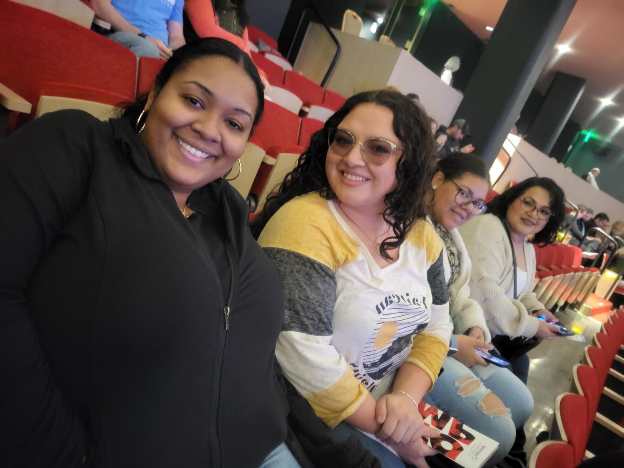 Four women seated in a theater with red seats, smiling at the camera.